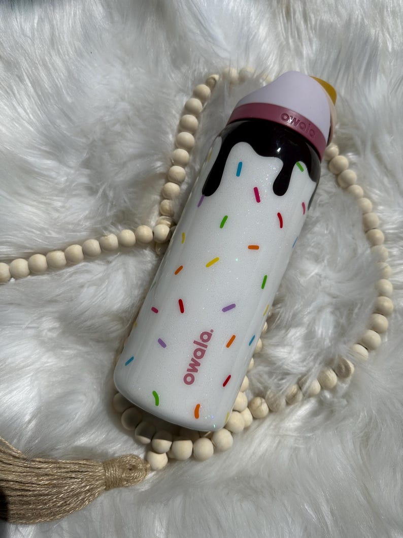 Puede incluir: Botella de agua reutilizable blanca con tapa rosa y un dise&ntilde;o marr&oacute;n parecido al chocolate con chispas de colores. La botella tiene la palabra "owala." impresa en ella.