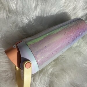 May include: A white and pink insulated tumbler with a glittery finish and a peach-colored lid. The tumbler has a cream-colored handle.