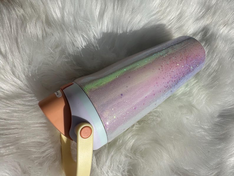 May include: A white and pink insulated tumbler with a glittery finish and a peach-colored lid. The tumbler has a cream-colored handle.