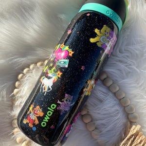 May include: Black Owala water bottle with a glittery finish, decorated with colorful cartoon characters, stars, and a unicorn. The bottle has a black lid with a teal handle and the brand name "owala" in green.