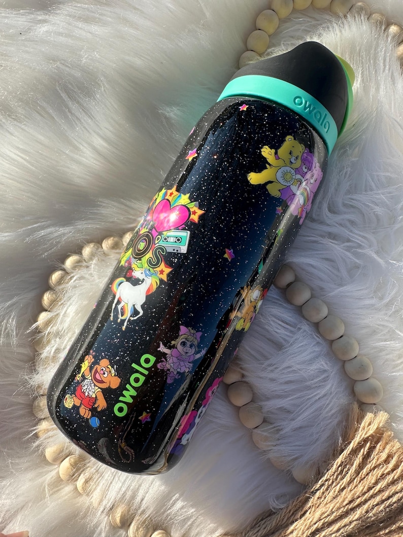May include: Black Owala water bottle with a glittery finish, decorated with colorful cartoon characters, stars, and a unicorn. The bottle has a black lid with a teal handle and the brand name "owala" in green.