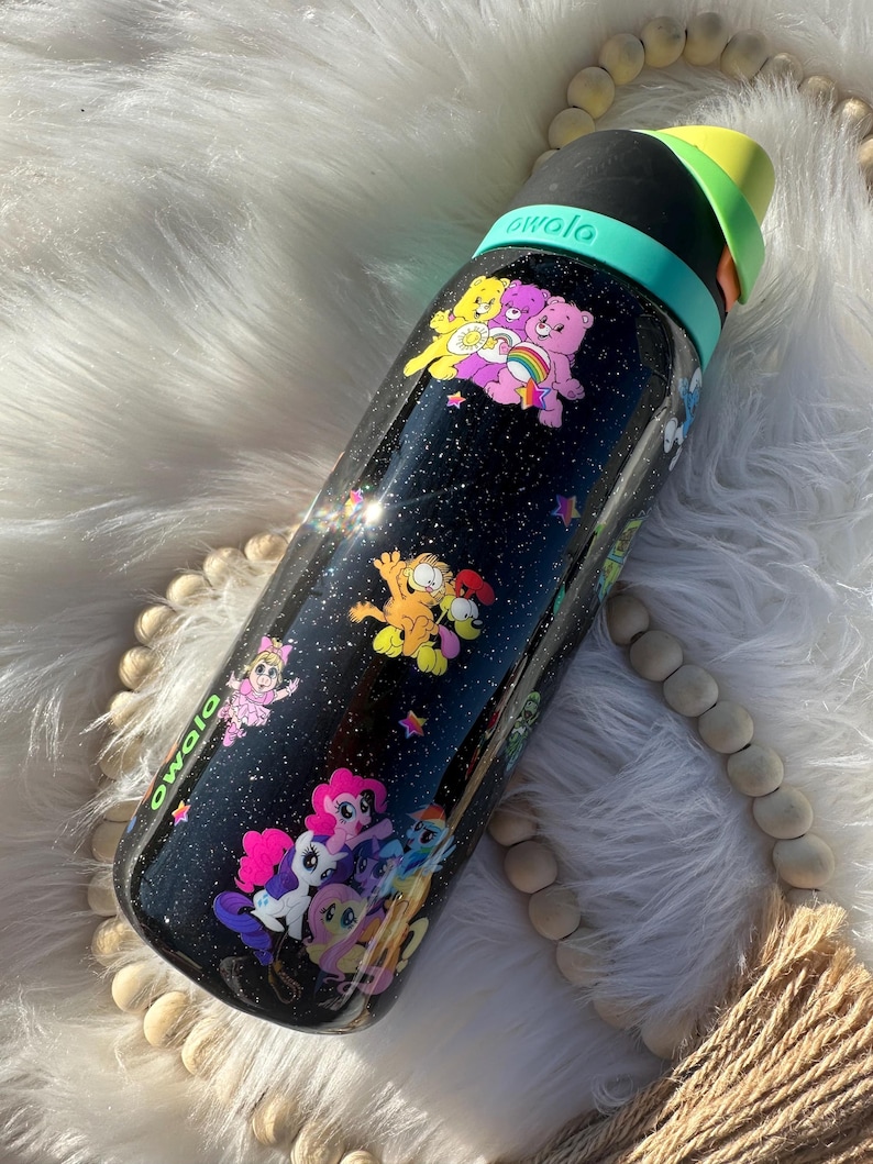 May include: Black Owala water bottle with a galaxy-themed design, featuring cartoon characters like Care Bears, Garfield, and My Little Pony. The bottle has a black lid with a green and yellow handle.