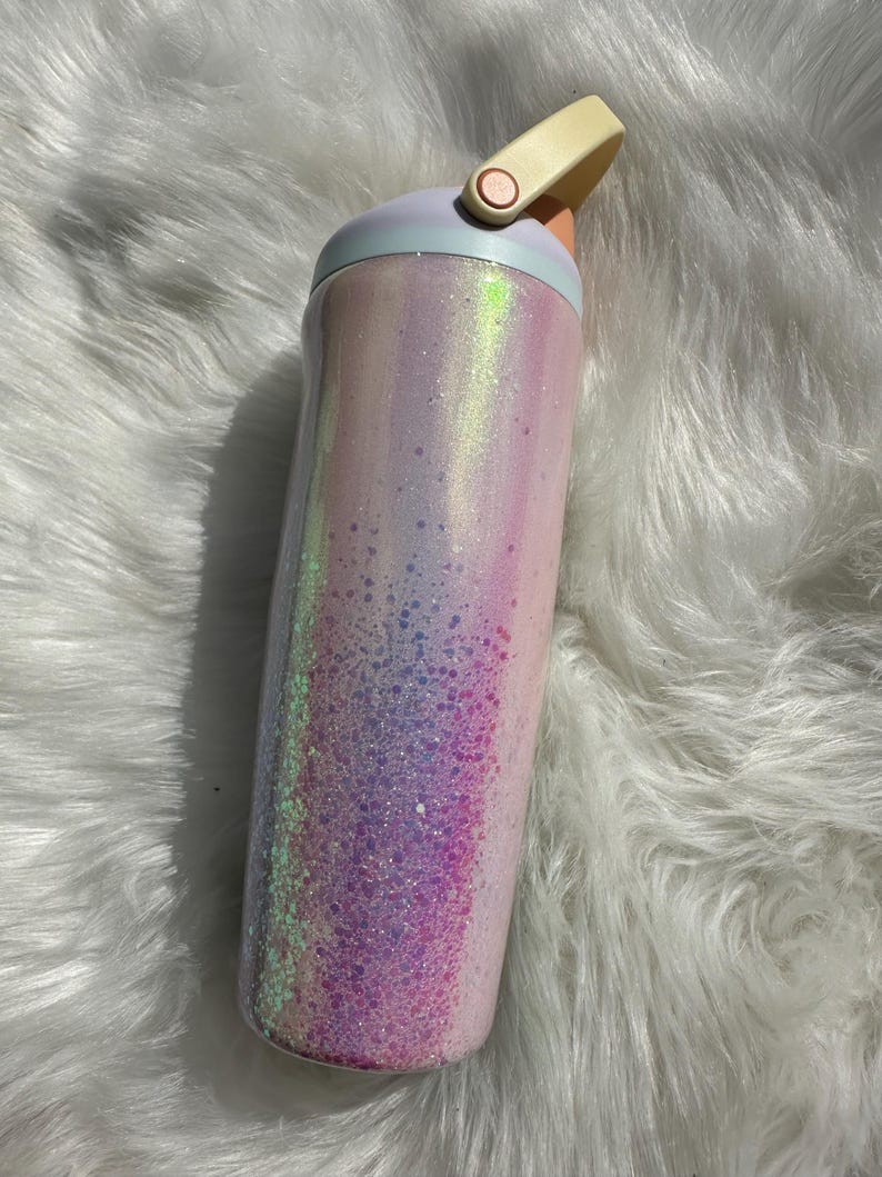 May include: A pink and white iridescent glitter tumbler with a light blue lid and a beige handle.
