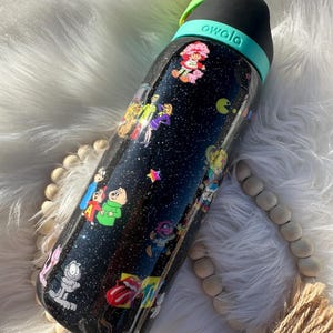 May include: Black Owala water bottle with a black lid and teal handle. The bottle is covered in colorful cartoon character stickers, including Strawberry Shortcake, Garfield, and Pac-Man. The bottle has a glittery finish.