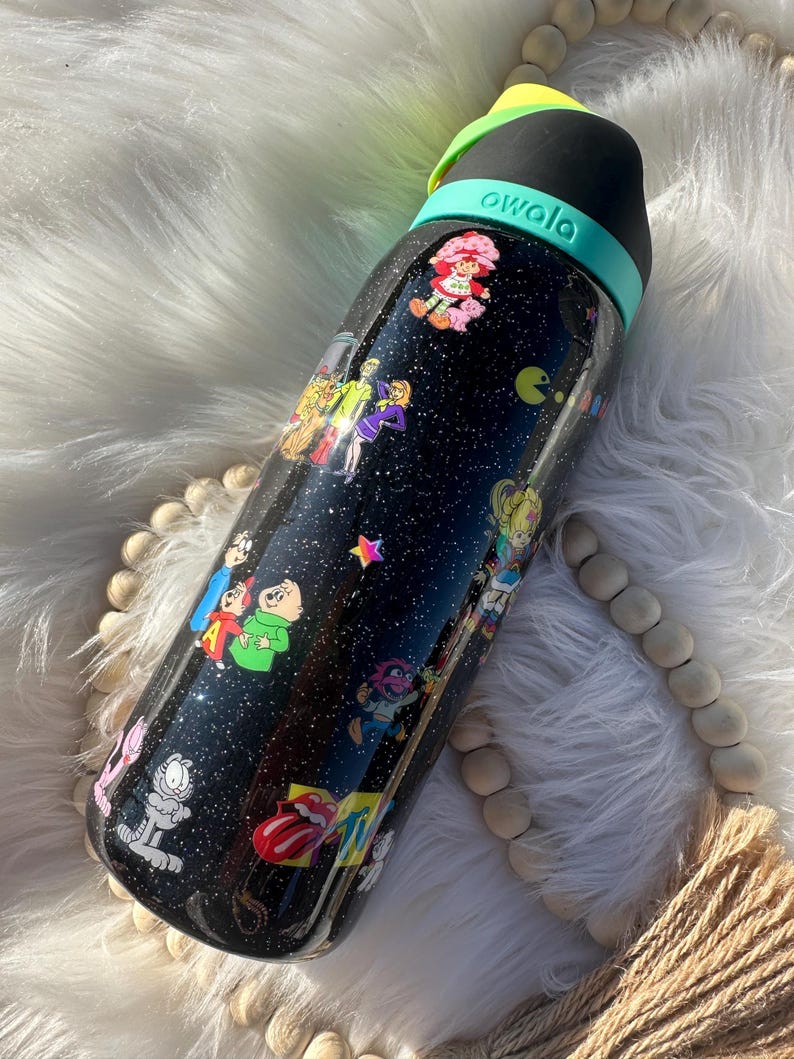 May include: Black Owala water bottle with a black lid and teal handle. The bottle is covered in colorful cartoon character stickers, including Strawberry Shortcake, Garfield, and Pac-Man. The bottle has a glittery finish.