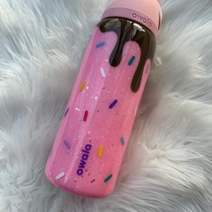 May include: Pink glitter water bottle with a chocolate drizzle design and colourful sprinkle accents. The bottle has a pink lid and the brand name "owala" is printed on the side. A fun and decorative water bottle.