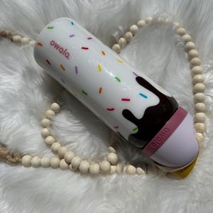 Puede incluir: Un vaso aislante blanco con un dise&ntilde;o de remolino de chocolate y blanco y chispas de colores. El vaso tiene una tapa rosa con la palabra "owala" impresa en &eacute;l.
