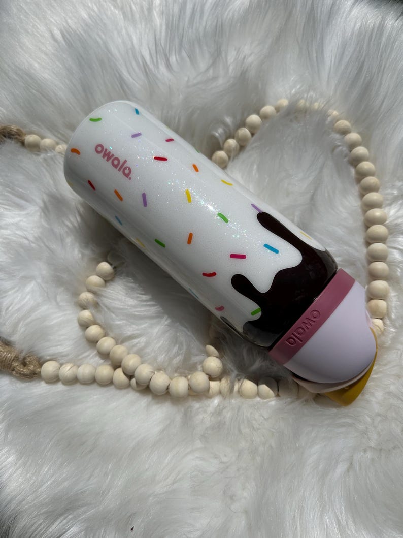 Puede incluir: Un vaso aislante blanco con un dise&ntilde;o de remolino de chocolate y blanco y chispas de colores. El vaso tiene una tapa rosa con la palabra "owala" impresa en &eacute;l.