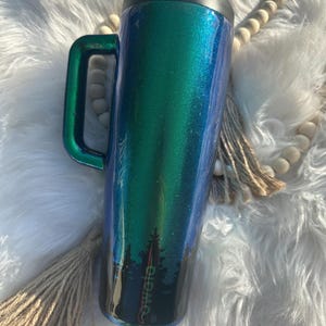 Peut inclure: Un gobelet isotherme avec une poignée, présentant un dégradé de couleurs bleu et vert. Le gobelet a un couvercle noir avec un bouton poussoir et une silhouette de forêt en bas. La marque "owala" est visible.