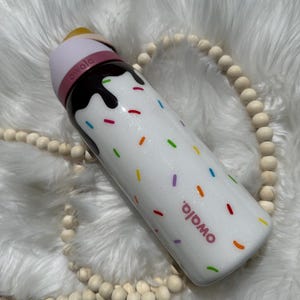 Puede incluir: Botella de agua reutilizable blanca con tapa rosa y un dise&ntilde;o marr&oacute;n parecido al chocolate con chispas de colores. La botella tiene la palabra "Omoio" impresa.