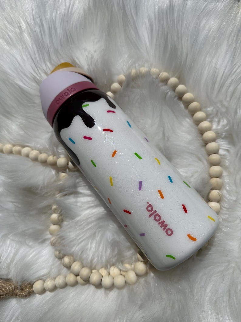 Puede incluir: Botella de agua reutilizable blanca con tapa rosa y un dise&ntilde;o marr&oacute;n parecido al chocolate con chispas de colores. La botella tiene la palabra "Omoio" impresa.