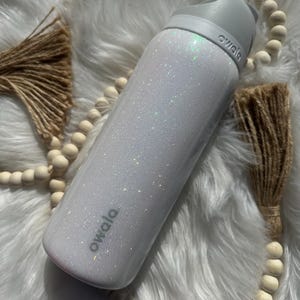 Puede incluir: Botella de agua reutilizable blanca con acabado de purpurina y la palabra "owala" impresa en el lateral.