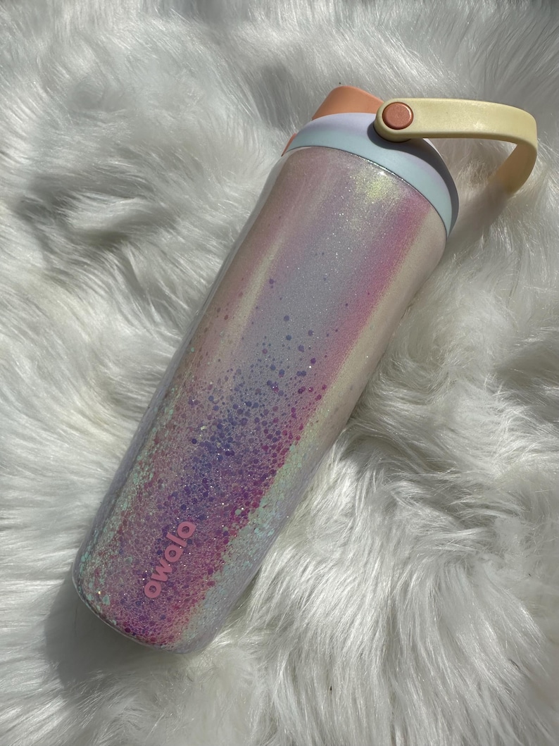 May include: A pink and white iridescent water bottle with a glitter finish and a yellow handle. The bottle has the word "owala" printed on the side.