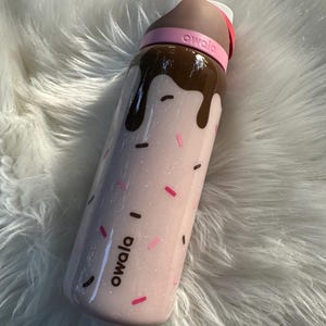 Puede incluir: Una botella de agua rosa Owala con un dise&ntilde;o de chocolate goteando por la parte superior. La botella tiene chispas rosas y negras y la palabra "owala". La tapa es marr&oacute;n y rosa.