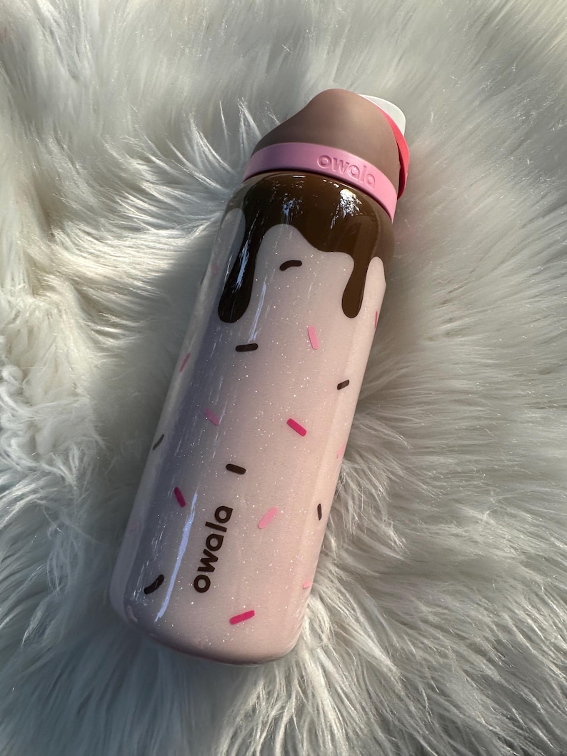 Puede incluir: Una botella de agua rosa Owala con un dise&ntilde;o de chocolate goteando por la parte superior. La botella tiene chispas rosas y negras y la palabra "owala". La tapa es marr&oacute;n y rosa.