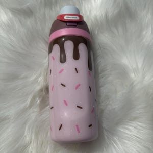 Puede incluir: Una botella de agua aislada rosa con un dise&ntilde;o caprichoso. La botella presenta una base rosa claro con chispas rosas y marrones, y goteo color chocolate. La parte superior tiene una tapa blanca y un bot&oacute;n rojo que dice "Lift to Unlock".