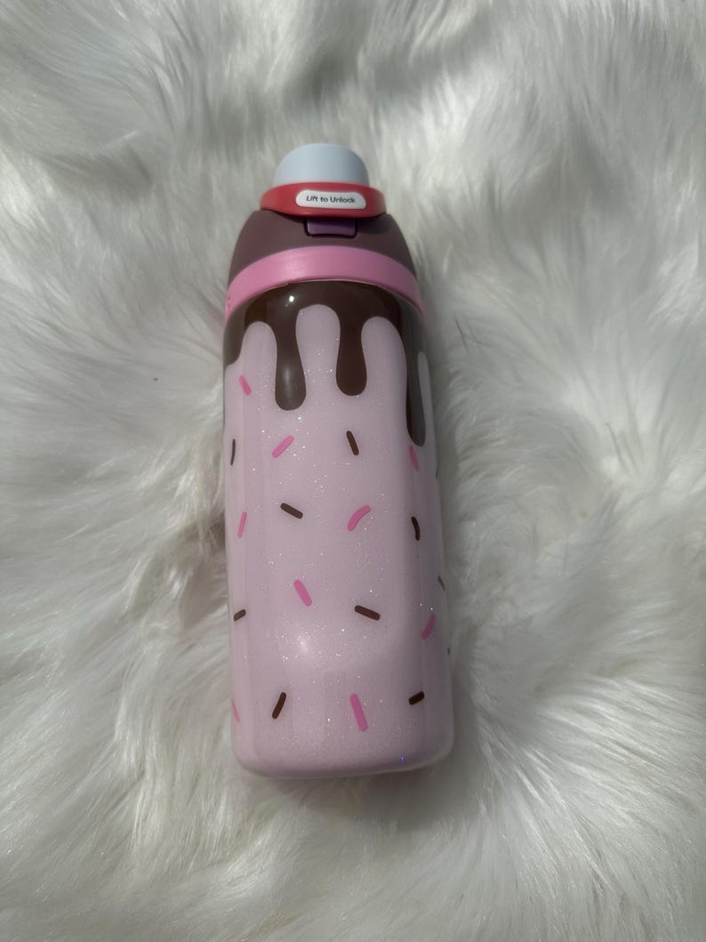 Puede incluir: Una botella de agua aislada rosa con un dise&ntilde;o caprichoso. La botella presenta una base rosa claro con chispas rosas y marrones, y goteo color chocolate. La parte superior tiene una tapa blanca y un bot&oacute;n rojo que dice "Lift to Unlock".
