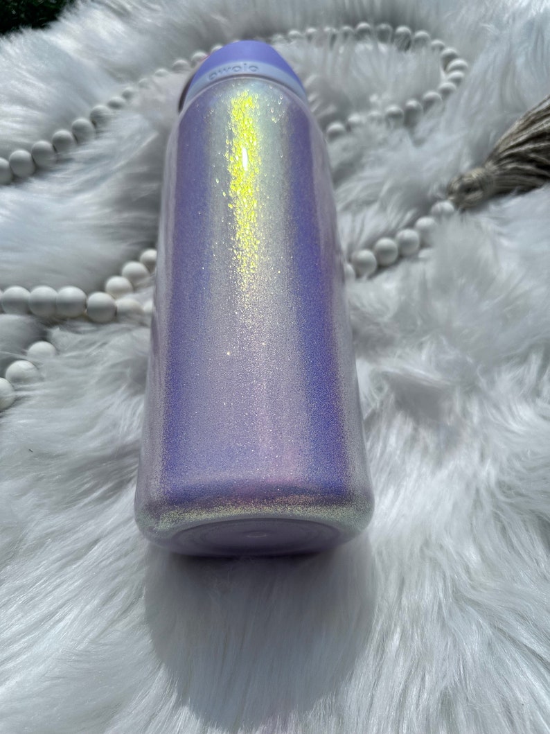 Op de afbeelding: Een paarse en witte glitterbeker met een lichtgele, iriserende glans. De beker heeft een schroefdop en een brede opening.