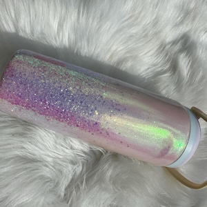 May include: A pink and purple glitter tumbler with a white lid and a beige handle. The tumbler has a holographic finish and is covered in iridescent glitter.