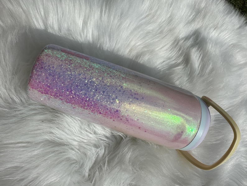 May include: A pink and purple glitter tumbler with a white lid and a beige handle. The tumbler has a holographic finish and is covered in iridescent glitter.