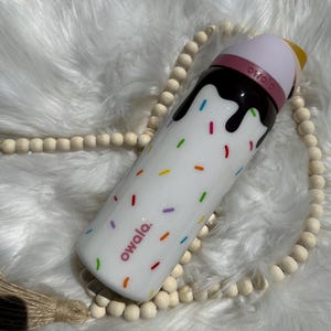 Puede incluir: Botella de agua reutilizable blanca con tapa rosa y marr&oacute;n. La botella tiene un dise&ntilde;o de chispas de colores y el texto "owala" impreso en ella.