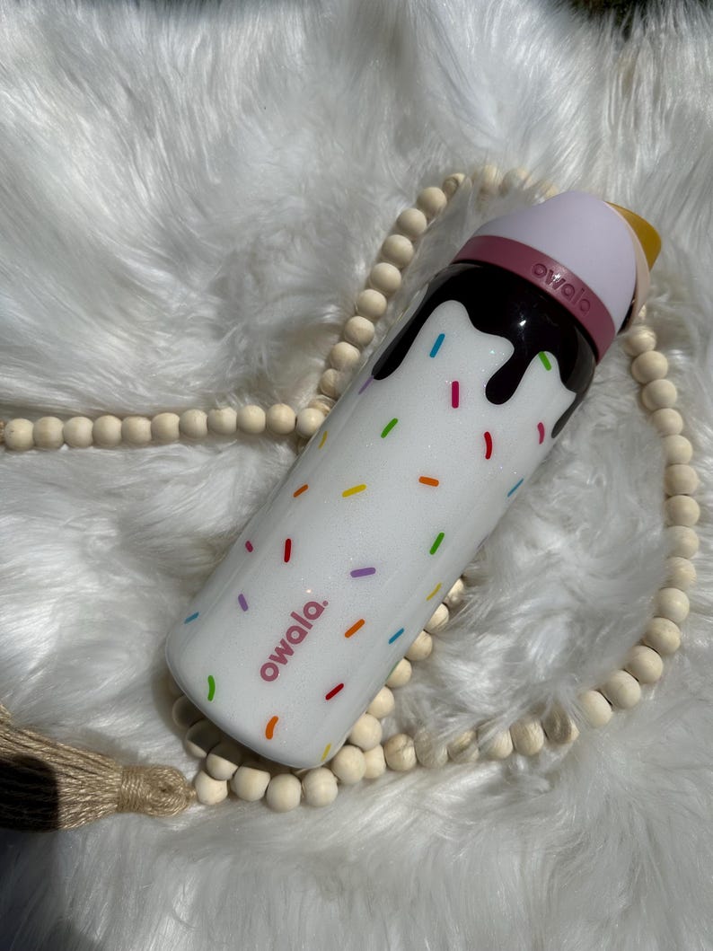 Puede incluir: Botella de agua reutilizable blanca con tapa rosa y marr&oacute;n. La botella tiene un dise&ntilde;o de chispas de colores y el texto "owala" impreso en ella.