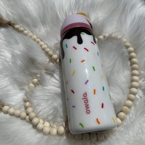 Puede incluir: Botella de agua aislada blanca con tapa rosa y dise&ntilde;o de goteo de chocolate. La botella tiene chispas de colores y la palabra "owala" impresa en ella.