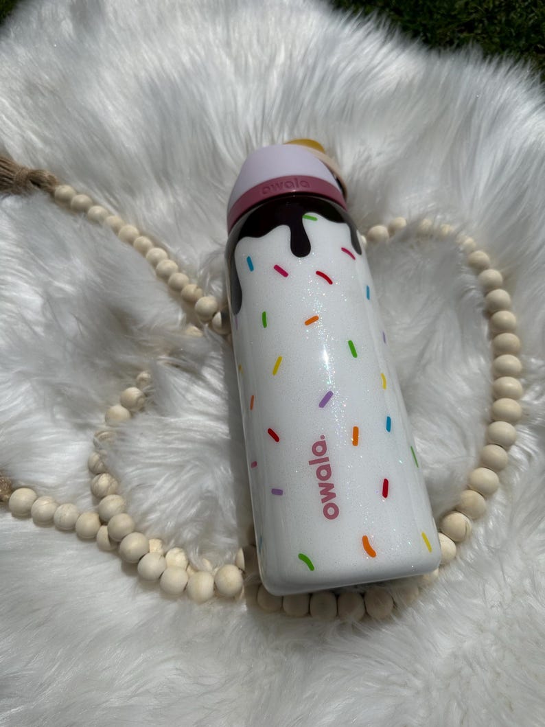 Puede incluir: Botella de agua aislada blanca con tapa rosa y dise&ntilde;o de goteo de chocolate. La botella tiene chispas de colores y la palabra "owala" impresa en ella.