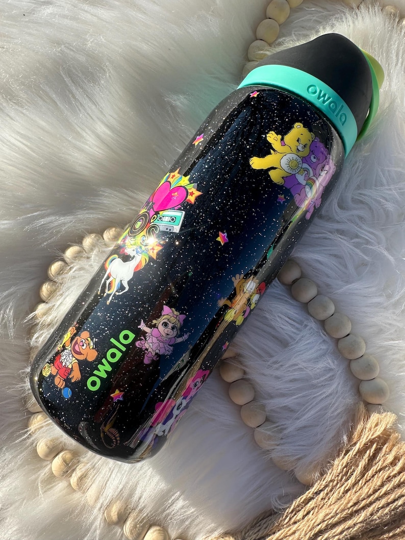May include: Black Owala water bottle with a variety of cartoon characters and a starry night design. The bottle features a black lid, a teal handle, and the Owala logo in green. The bottle is decorated with Care Bears, unicorns, and other colorful graphics.