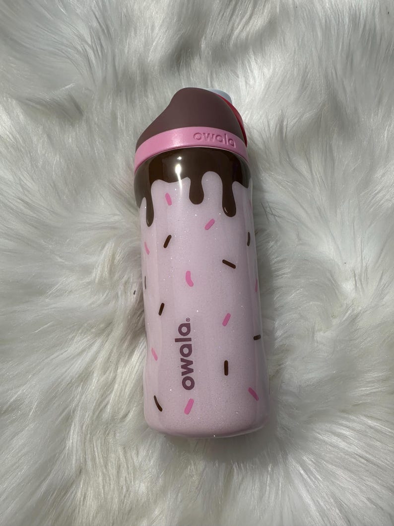 Puede incluir: Una botella de agua rosa Owala con tapa marr&oacute;n y banda rosa. La botella tiene un dise&ntilde;o de chispas rosas y gotas marrones. El logotipo de Owala est&aacute; impreso en el lateral. La botella est&aacute; dise&ntilde;ada para parecerse a un postre.