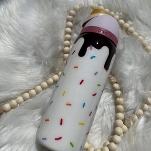 Puede incluir: Botella de agua aislada blanca con un dise&ntilde;o marr&oacute;n parecido al chocolate y chispas de colores. La botella tiene una tapa rosa con la palabra "owala" en ella.