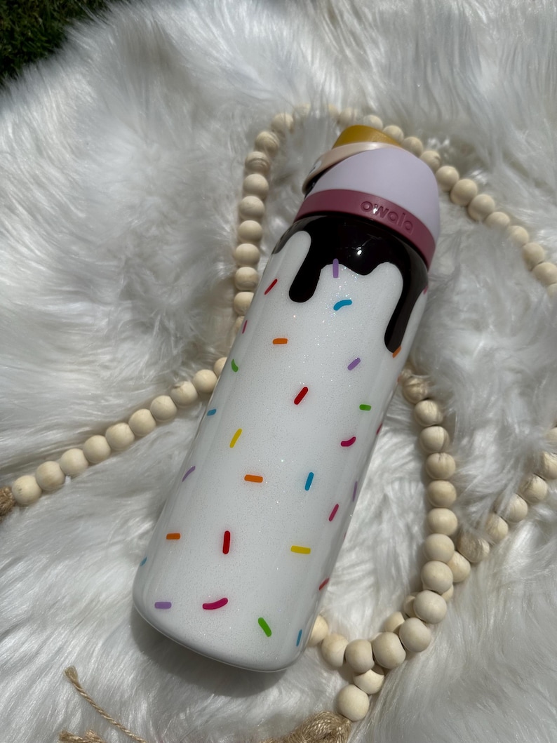 Puede incluir: Botella de agua aislada blanca con un dise&ntilde;o marr&oacute;n parecido al chocolate y chispas de colores. La botella tiene una tapa rosa con la palabra "owala" en ella.