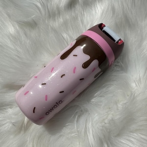 Puede incluir: Una botella de agua aislada rosa Owala con un dise&ntilde;o de goteo de chocolate y chispas. La botella tiene una tapa marr&oacute;n y rosa y la palabra "owala" est&aacute; impresa en el lateral.