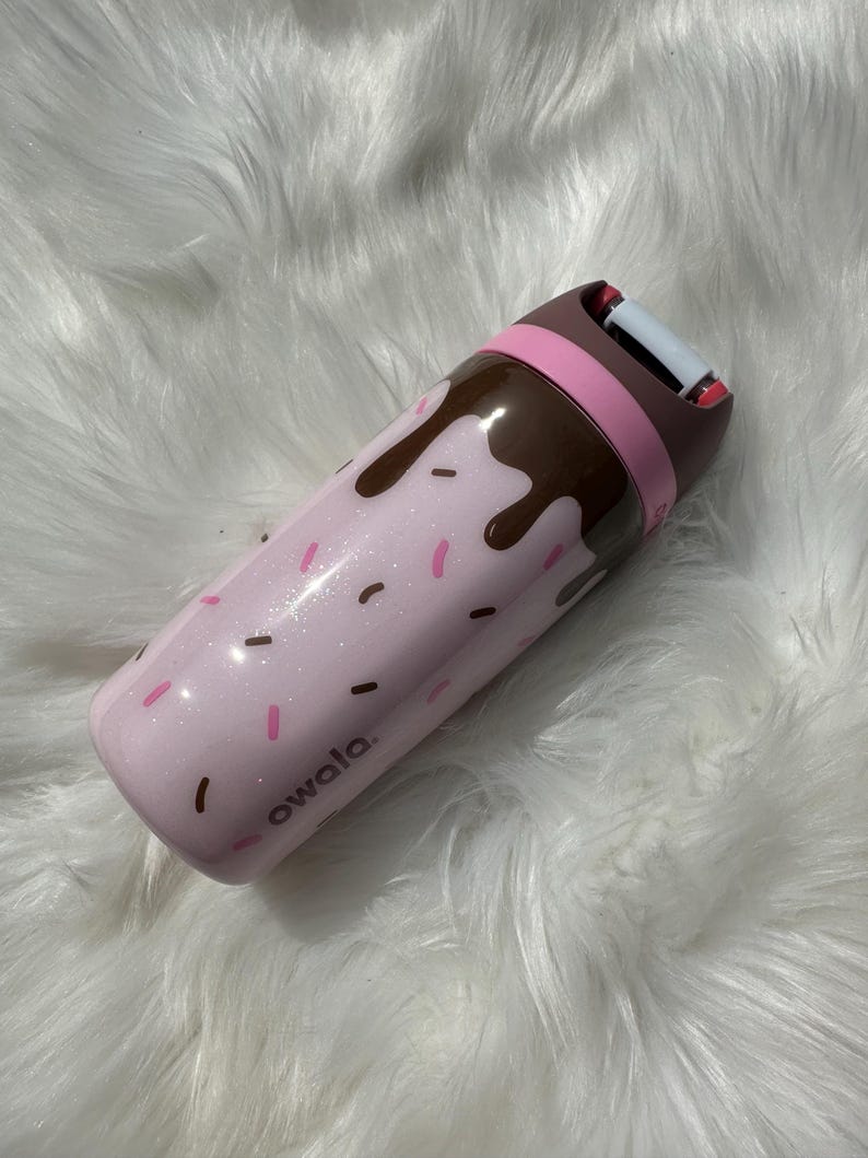 Puede incluir: Una botella de agua aislada rosa Owala con un dise&ntilde;o de goteo de chocolate y chispas. La botella tiene una tapa marr&oacute;n y rosa y la palabra "owala" est&aacute; impresa en el lateral.