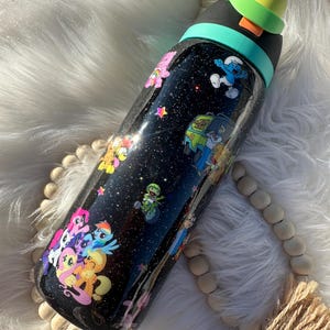 May include: A black water bottle with a glittery finish, adorned with colorful cartoon characters like My Little Pony, the Smurfs, and Scooby-Doo. The bottle has a black lid with a yellow cap and a teal band.