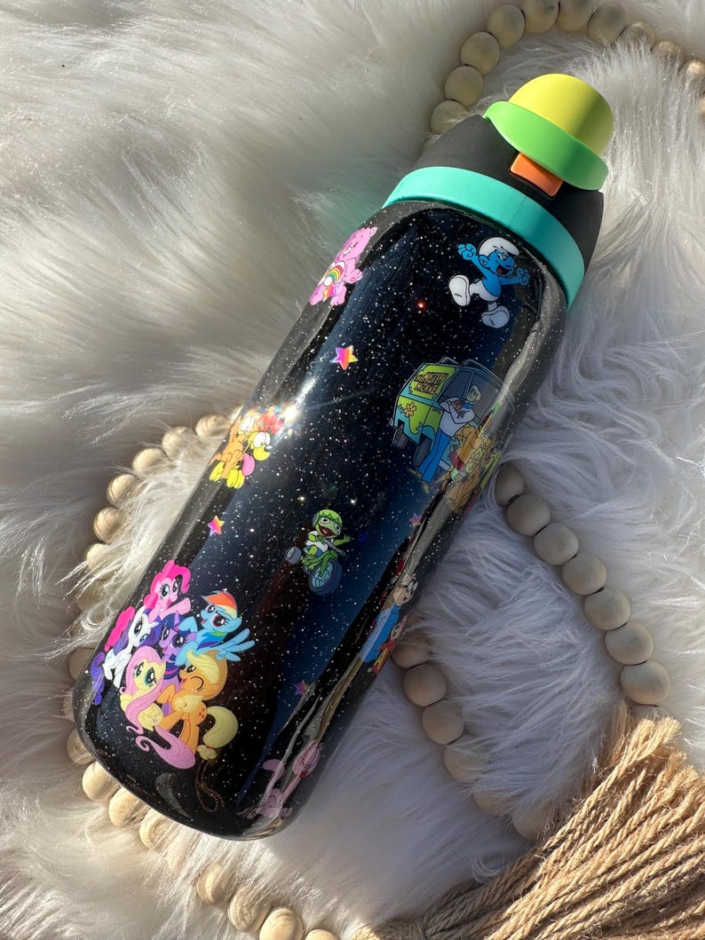 May include: A black water bottle with a glittery finish, adorned with colorful cartoon characters like My Little Pony, the Smurfs, and Scooby-Doo. The bottle has a black lid with a yellow cap and a teal band.