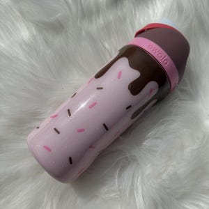 Puede incluir: Una botella de agua rosa con un dise&ntilde;o de goteo de chocolate y chispas rosas y marrones. La botella tiene una tapa marr&oacute;n y una banda rosa con la palabra "owala" impresa.