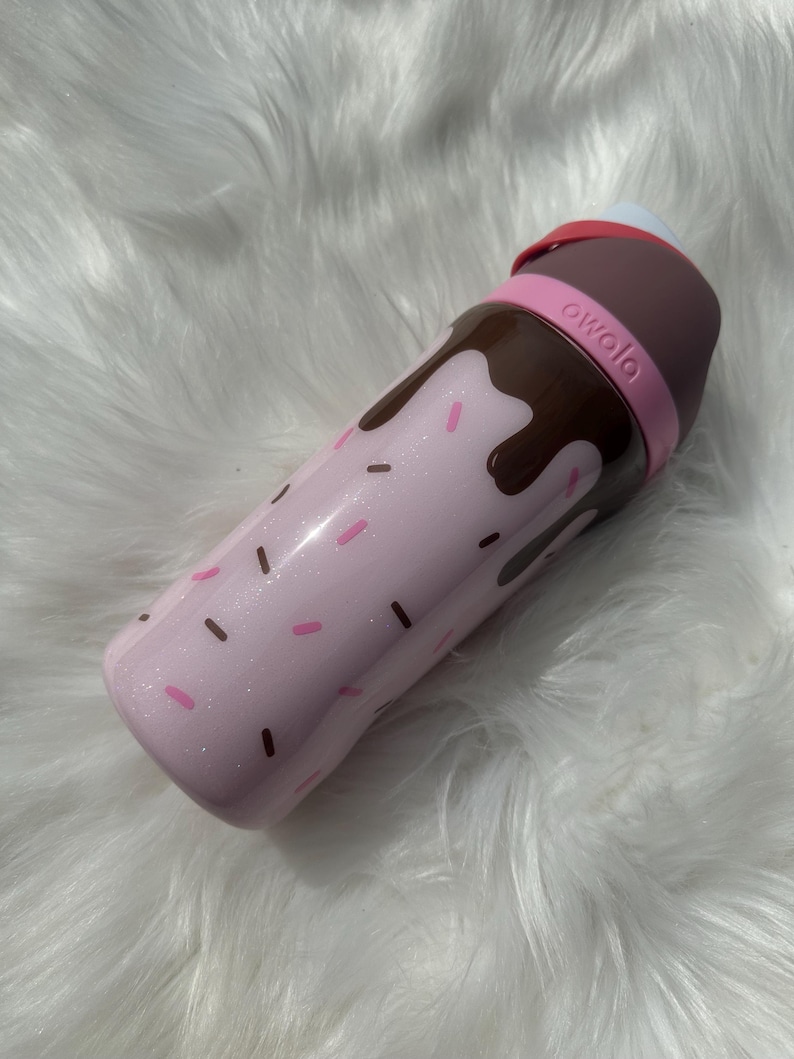 Puede incluir: Una botella de agua rosa con un dise&ntilde;o de goteo de chocolate y chispas rosas y marrones. La botella tiene una tapa marr&oacute;n y una banda rosa con la palabra "owala" impresa.