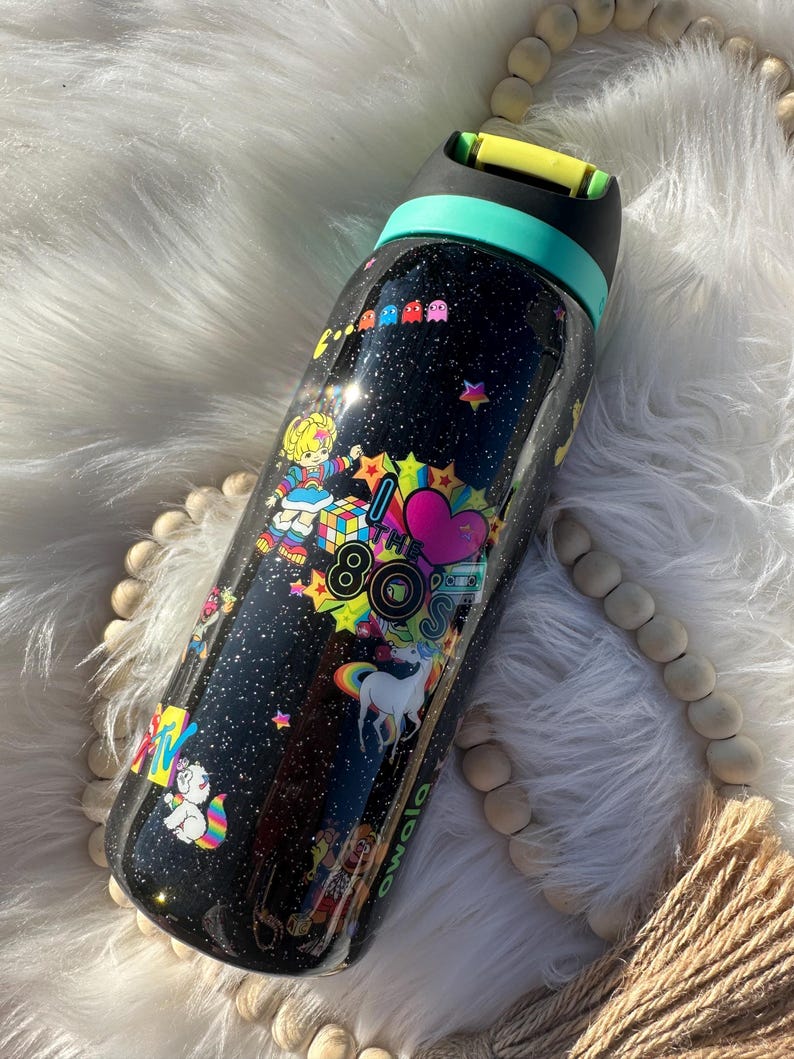 May include: Black water bottle with a glittery finish, decorated with colorful 80s-themed graphics. The design includes a unicorn, Pac-Man characters, and the text "80s". The bottle has a teal and yellow lid.