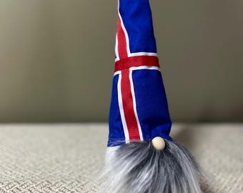 May include: A decorative gnome with a blue hat featuring the Icelandic flag design. The hat has a red band and white stripes. The gnome has a long, gray and white beard and a small wooden nose. The gnome is a festive decoration.