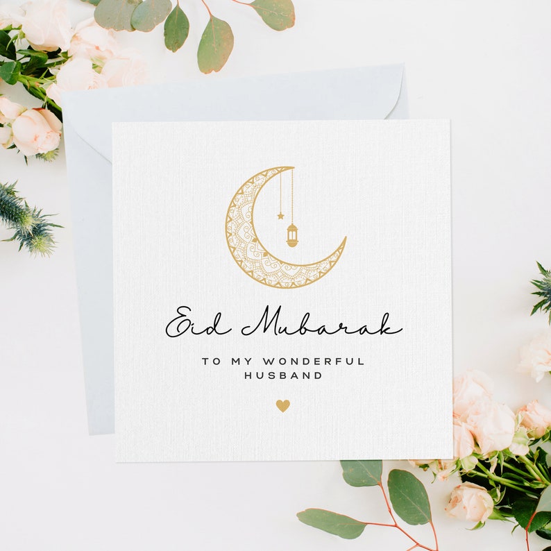 May include: A white card with a gold crescent moon and a hanging lantern design. The text "Eid Mubarak" is written in black cursive script. Below the text is "To my wonderful husband" with a small heart symbol.
