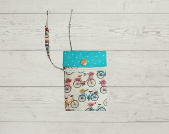 May include: A small crossbody bag featuring a cream-coloured fabric with a repeating pattern of colourful bicycles carrying flower baskets. The bag has a bright turquoise flap with white polka dots, a circular floral button, and a matching fabric strap.