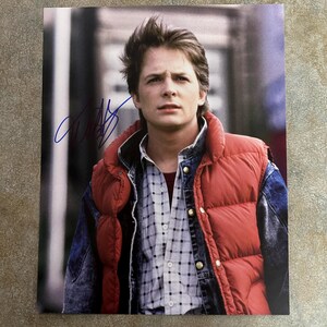May include: A black and white photo of a young man wearing a red puffer vest over a blue denim jacket and a plaid shirt. The photo is signed with a blue signature.