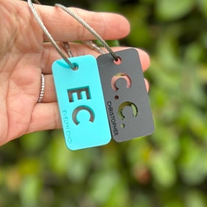 May include: Two turquoise and black luggage tags with metal rings. The turquoise tag has the initials "EC" and the name "Evelyn Choi" printed on it. The black tag has the initials "C.C." and the name "Christopher" printed on it.