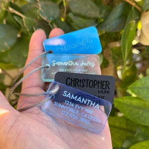 May include: Three clear acrylic keychains with metal rings. The top keychain is light blue and says "Samantha Broker". The middle keychain is clear and says "Samantha Jules 321 Evylittle Way". The bottom keychain is clear and says "Samantha Evylittle Way 1234 Evy Way Sunnyvale, CA 01234 123-456-7890". The bottom keychain also has a black acrylic keychain with the text "Christopher Muni" on it.