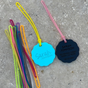 May include: Two round, personalized name tags with a blue and black background. The blue tag says "Sarah Johnson" and the black tag says "Sarah Johnson" with an address and phone number. A bundle of colorful rubber bands are in the background.