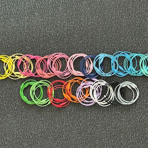 May include: Assortment of colorful elastic hair ties in various shades of pink, yellow, blue, green, orange, and purple. The hair ties are arranged in a row on a dark gray surface.