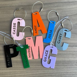 May include: A collection of colorful luggage tags in various shapes, each with a name printed on it. The tags are in shades of pink, orange, blue, green, purple, and black, with silver metal loops for attachment. Names include Cassie, Andy, Julia, Casey, Pablo, Trevor, Maddie, Grace.