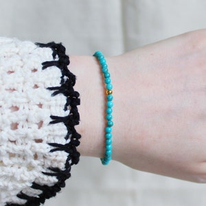 May include: A turquoise beaded bracelet with a gold bead accent. The bracelet is on a person's wrist.