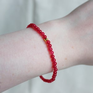 May include: A red beaded bracelet with gold accents. The bracelet is made of small, round beads and is worn on a person's wrist.